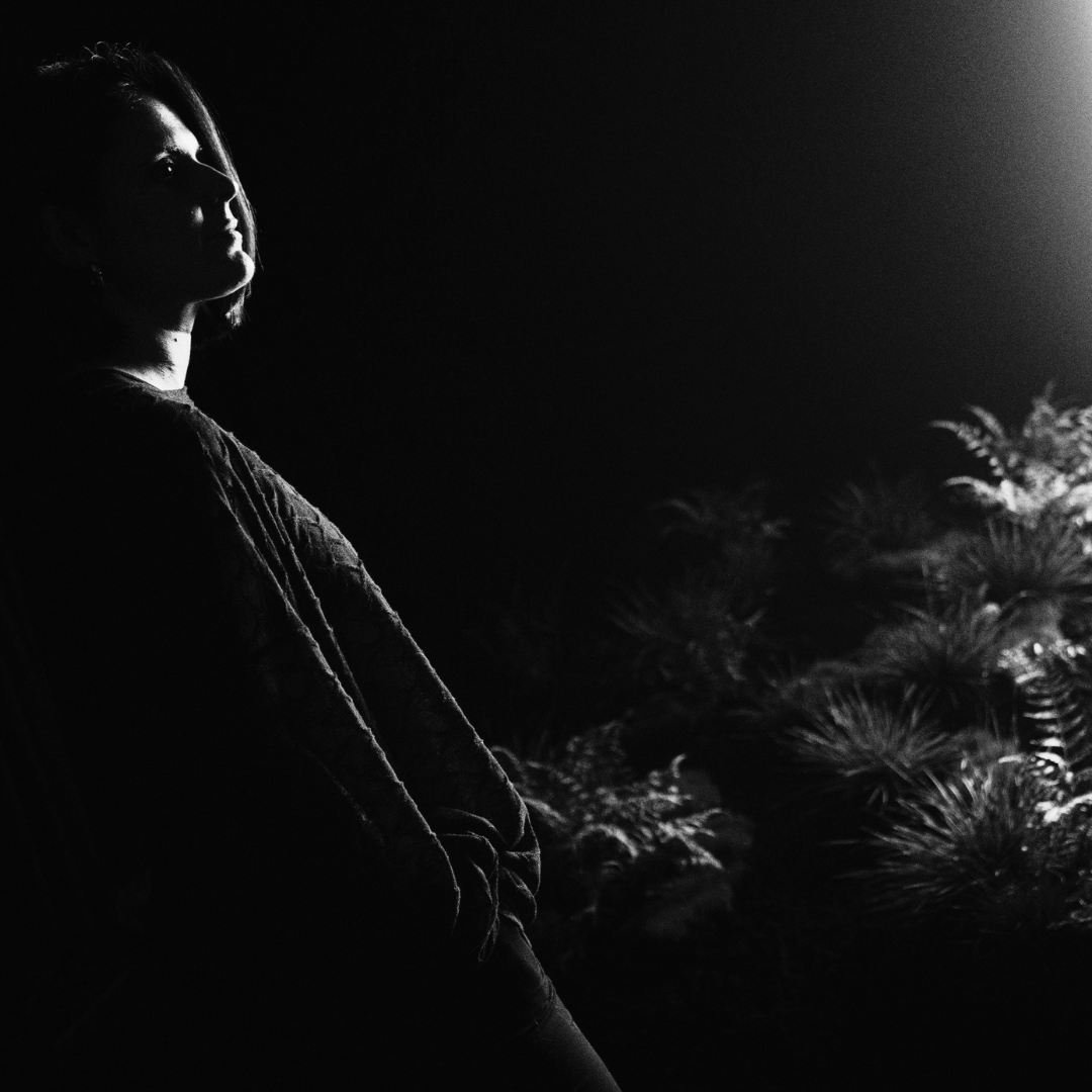 Image en noir et blanc de l'artiste Cinzia Campolese dans la pénombre près d'une plante verte à côté d'une fenêtre d'où émane de la lumière blanche. Pour la résidence de recherche du Chantier IA à Sporobole