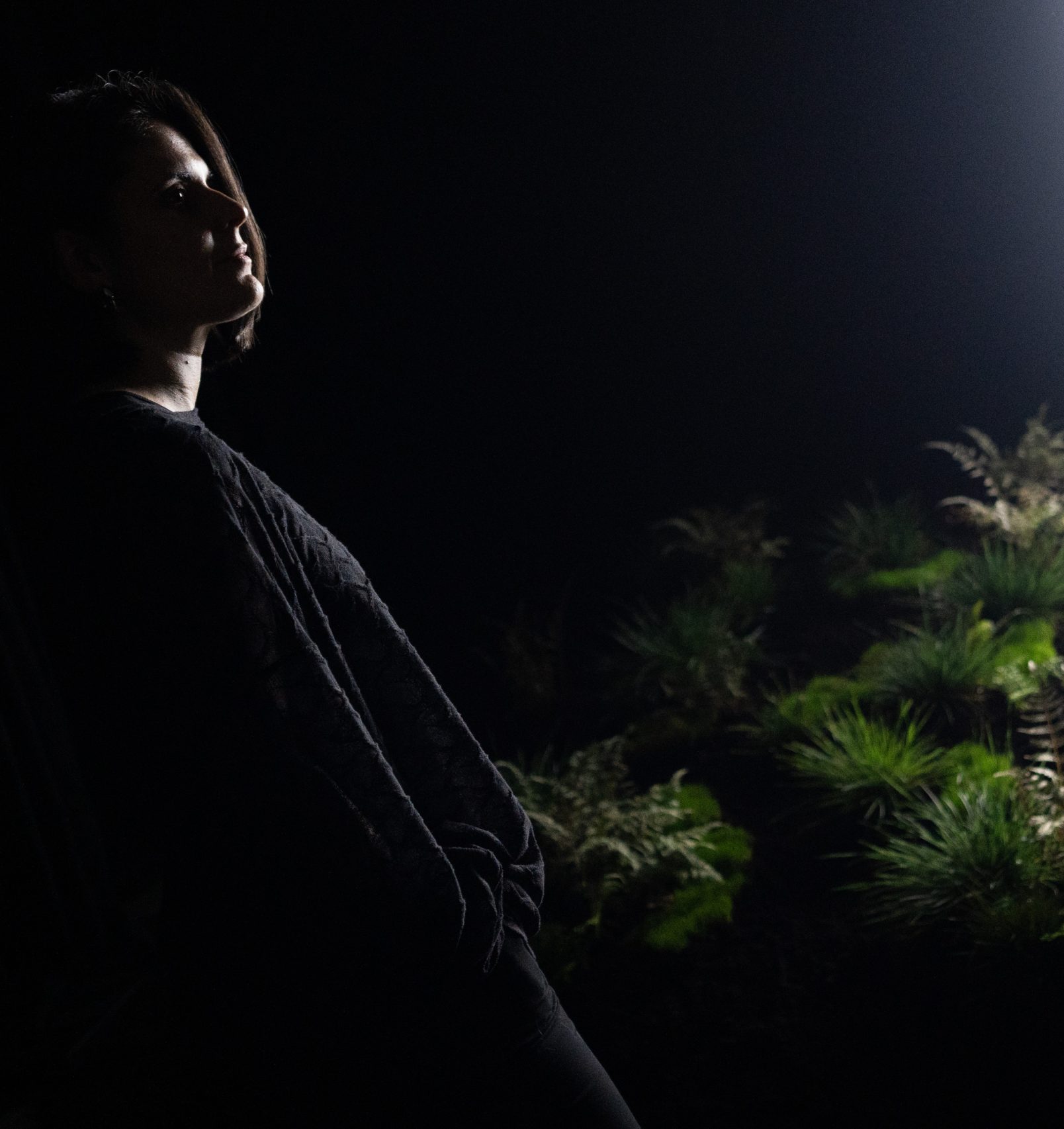 Image de l'artiste Cinzia Campolese dans la pénombre prè d'une plante verte à côté d'une fenêtre d'où émane de la lumière blanche. Pour la résidence de recherche du Chantier IA à Sporobole