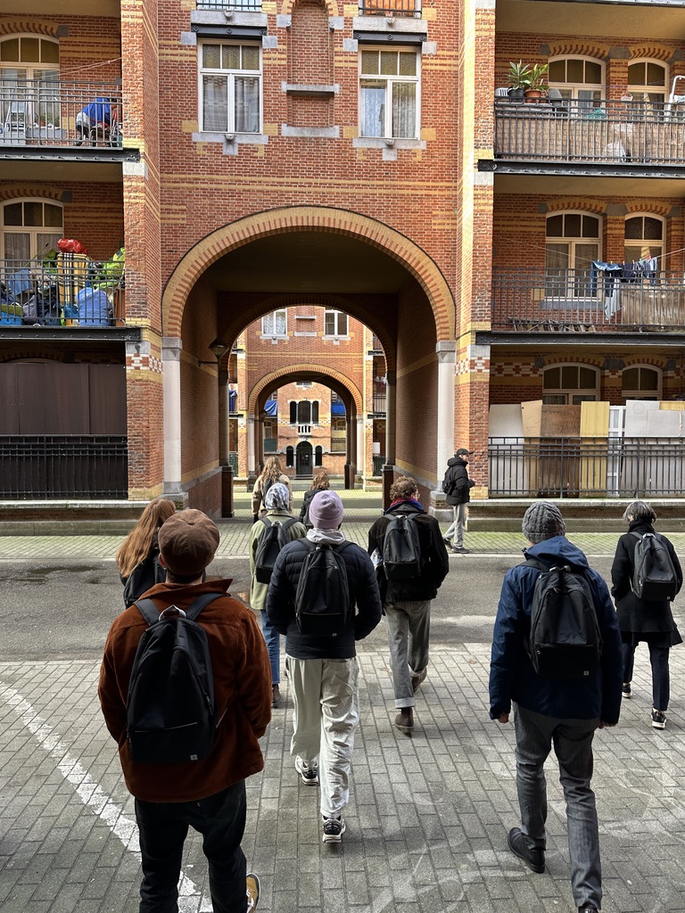 Photo d’un petit groupe de personnes marchant dans une cour entourée d’immeubles en briques rouges. Elles portent des manteaux d’hiver, des bonnets et des sacs à dos, et avancent sous une grande arche qui relie deux bâtiments. Les façades présentent des balcons avec des plantes et du linge, et une succession d’arcs en briques crée une perspective vers d’autres cours semblables à l’arrière-plan. La lumière est douce, probablement en fin de matinée ou par temps couvert. Ce sont des participantes et participants à l'oeuvre A Chorus of Footsteps d'aifoon pour le Mois pédagogique 2026 à Sporbole.