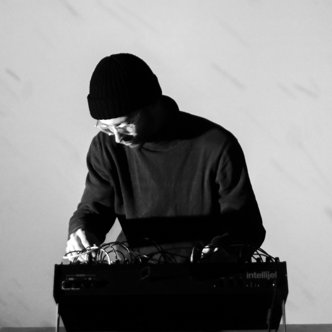 Photo en noir et blanc montrant un homme, l'artiste Jérémie Martineau, concentré manipulant un synthétiseur modulaire ou un boîtier électronique. Il porte un bonnet noir, des lunettes transparentes et un chandail sombre à col montant. Il est penché vers son instrument, les deux mains sur les câbles et boutons, dans une lumière douce qui met en valeur son visage et ses gestes. L’arrière-plan est uni et clair. Pour le Mois pédagogique 2026 de Sporobole.