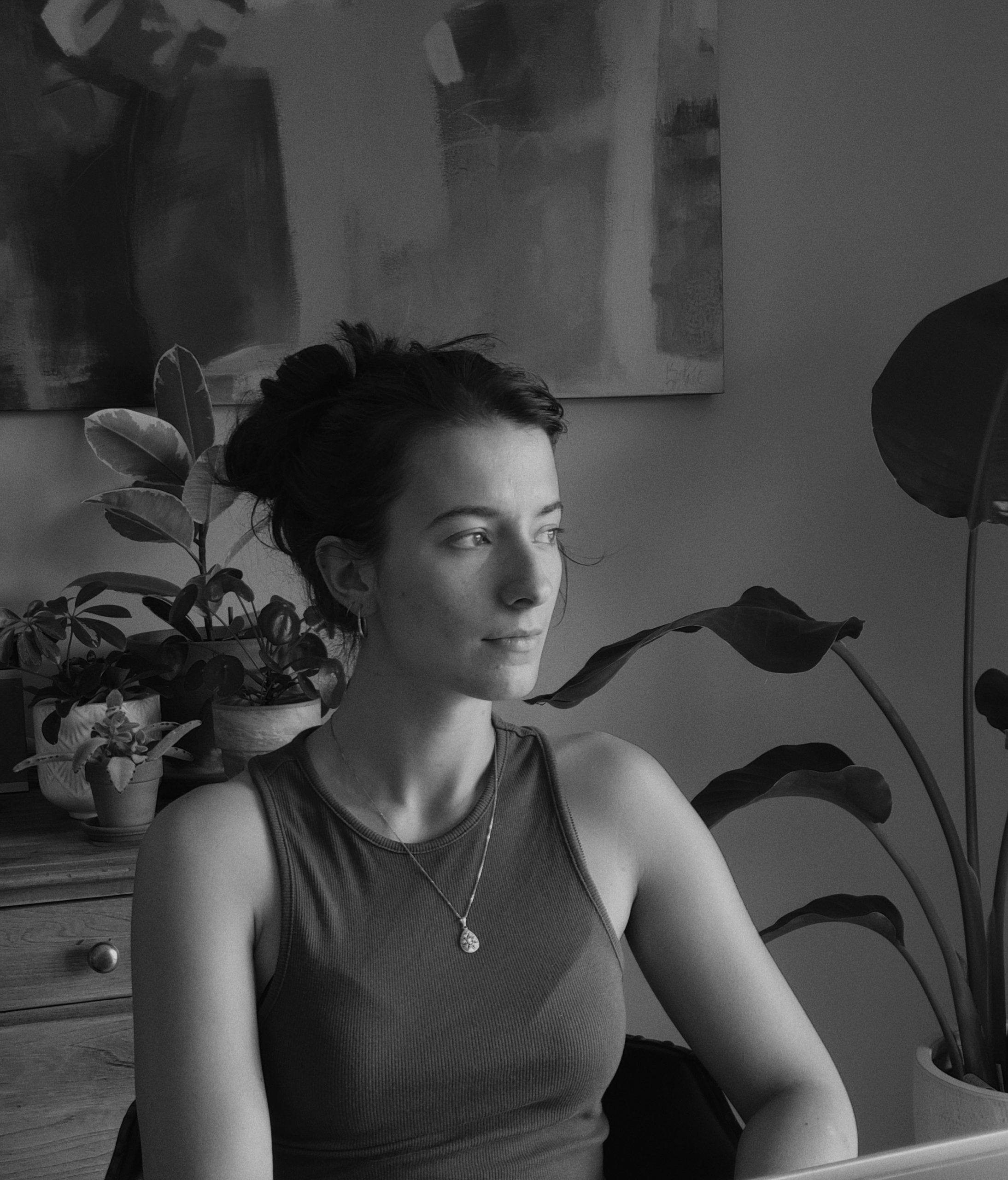 Portrait photographique en noir et blanc de Tamara Krpic, cadré à partir de la poitrine. Elle est légèrement de profil, le regard tourné sereinement vers la droite de l'image. Ses cheveux foncés sont relevés en un chignon décontracté, et elle porte de petites boucles d'oreilles ainsi qu'un collier à chaîne fine orné d'un pendentif en forme de goutte. Elle est vêtue d'un haut sans manches à encolure ronde. L'arrière-plan, un intérieur chaleureux et légèrement estompé, laisse transparaître plusieurs plantes vertes en pot, dont un caoutchouc à grandes feuilles sur la gauche, créant un cadre naturel et intime. Pour la résidence Fictions d'IA à Sporobole