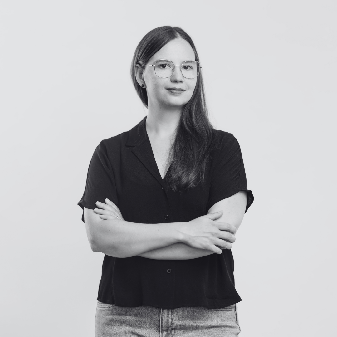 Portrait photo en noir et blanc de Christiane Vadnais. Une femme les bras croisés sur une chemise noire à manches courtes. Elle a les cheveux longs placés sur le côté droit et elle porte des lunettes et un jeans. Pour le Chantier IA de Sporobole. résidence Fictions d'IA.