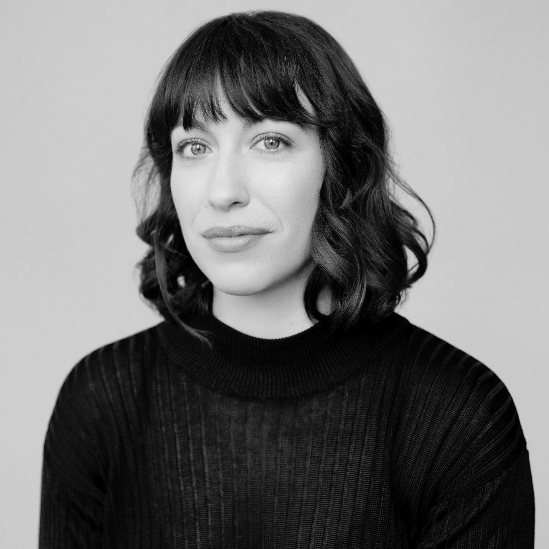 Portrait photographique de face et en noir et blanc de Maude Jarry, cadré à partir de la poitrine. Elle regarde l'objectif avec une expression douce et un léger sourire serein. Ses cheveux foncés sont mi-longs, ondulés et coupés avec une frange droite qui s'arrête juste au-dessus des sourcils. Elle porte un chandail noir à col montant, caractérisé par une texture de fines côtes verticales. Le fond derrière elle est entièrement uni, épuré et de couleur gris clair, mettant pleinement en valeur son visage. Pour la résidence de recherche Fictions d'IA à Sporobole
